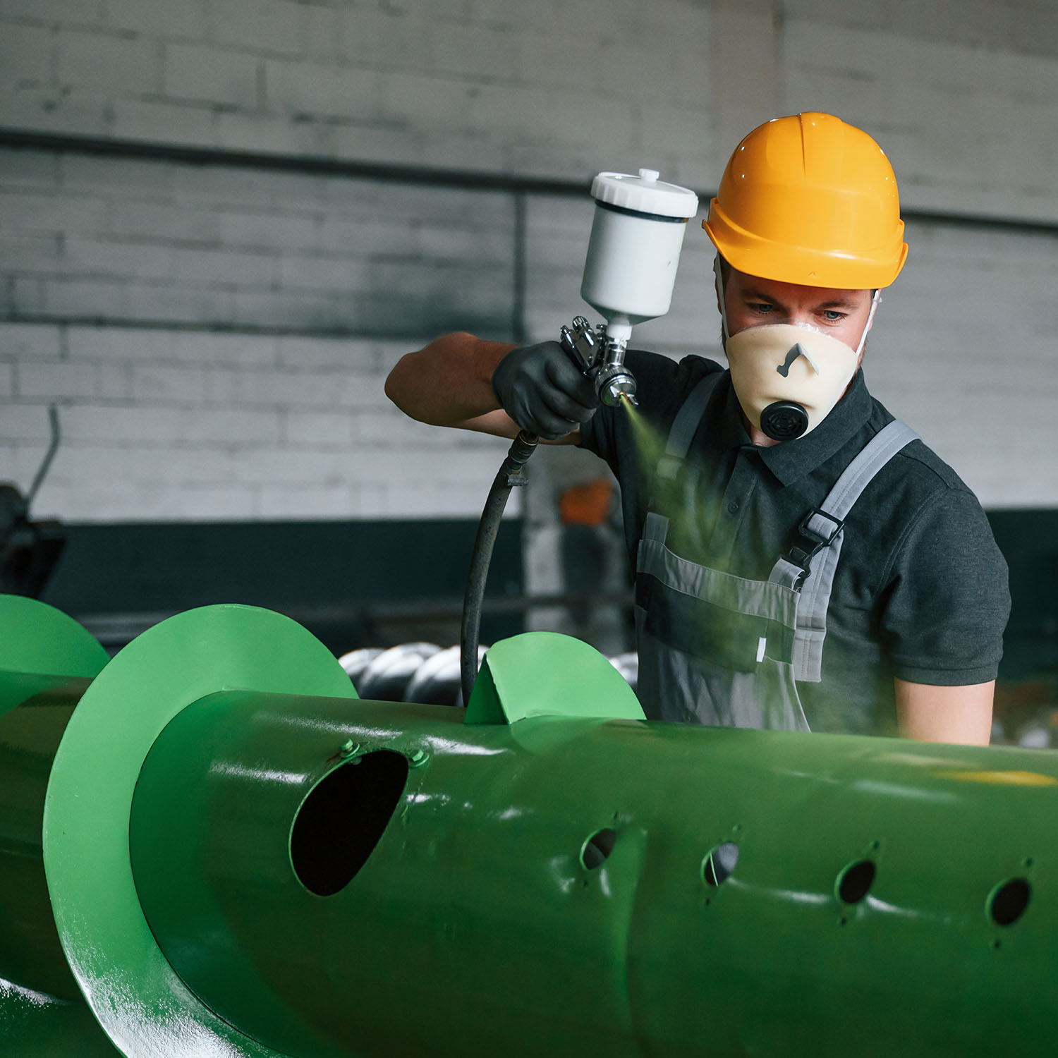 A man with PPE and a spray gun spraying some industrial metalwork in green paint.